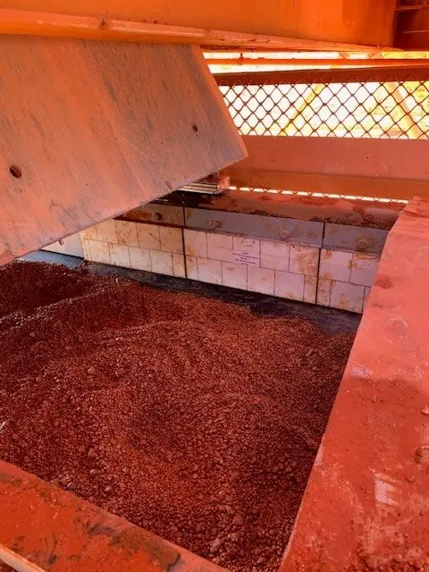 A close-up view of red ore or mineral material inside an industrial machine with metal grating and orange-tinted surfaces, partially covered by a hinged metal panel.