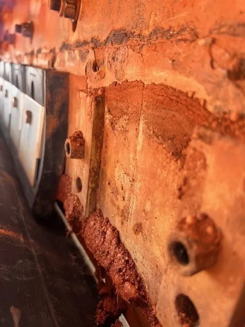 Close-up view of a rusty, corroded metal surface with bolts and reddish-brown rust buildup, possibly part of industrial machinery or a vehicle component. The image is focused on the textured, deteriorated metal.