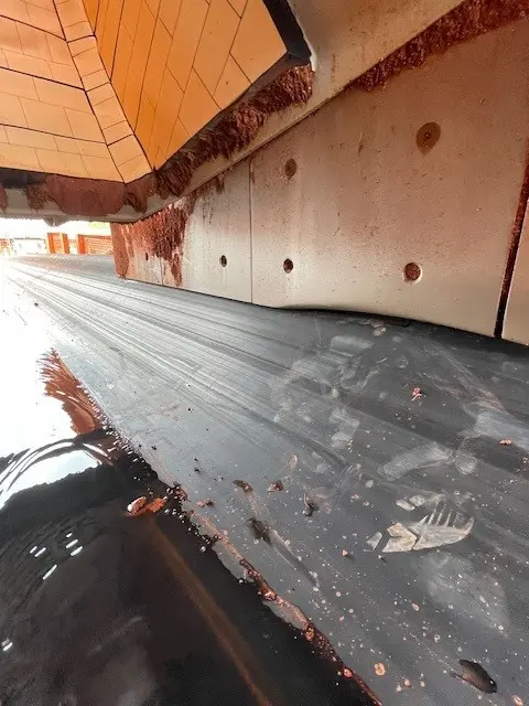 A conveyor belt with a black surface and scattered reddish-brown residue moves under a tiled chute and metal wall inside an industrial facility.