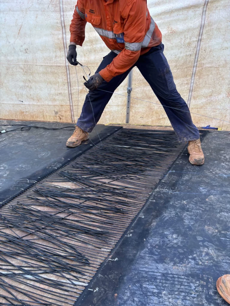 A worker in an orange shirt and blue pants stands on a black surface, handling long black wires laid out in parallel rows on a mat, inside a tent-like structure.