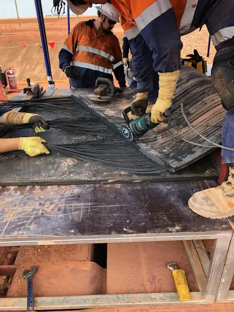 Workers in safety gear use power tools and gloves to cut and repair a damaged conveyor belt, with black cords exposed, at an industrial worksite. Tools and equipment are visible around them.