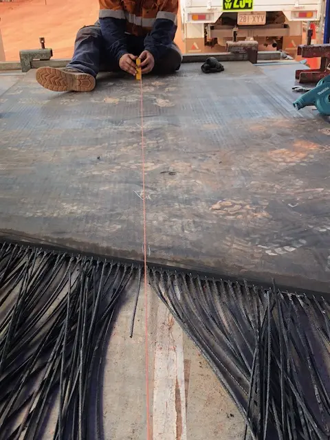 A person sitting on a flat surface pulls a measuring tape along an industrial rubber conveyor belt with cut rubber strips at the edge, likely preparing for installation or repair. A truck and dirt ground are visible in the background.