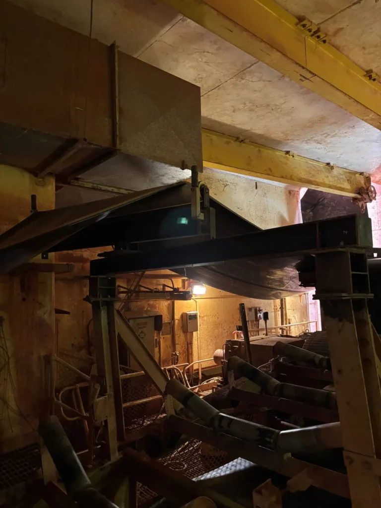 Large industrial conveyor system inside a dimly lit factory, with yellow beams, metal structures, pipes, and machinery visible in the background. A single light bulb illuminates part of the scene.