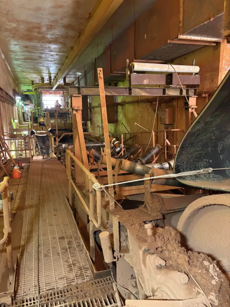 A dusty industrial walkway with metal railings runs alongside machinery and conveyor belts in a factory setting; a worker in safety gear is seen in the distance.
