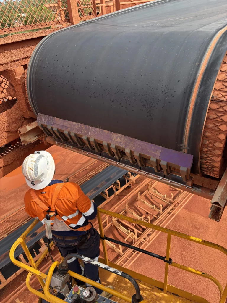 A worker in a white helmet and orange safety gear stands on a yellow platform, inspecting or maintaining a large black conveyor belt system in an industrial setting with red dust and metal structures.
