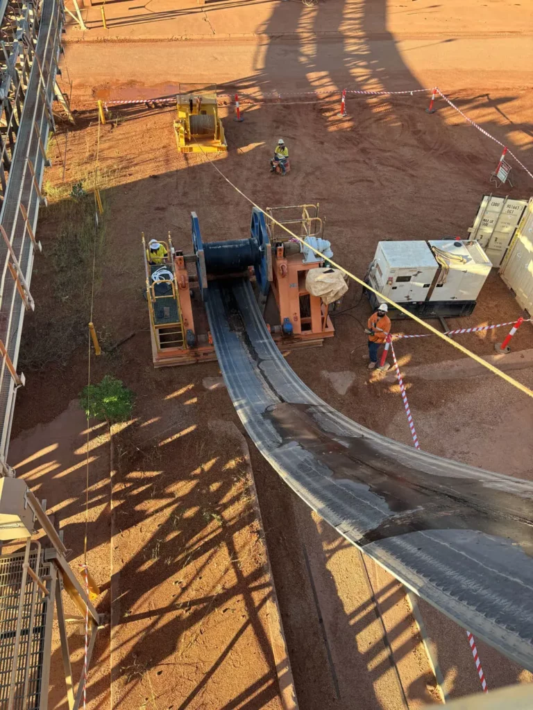 Aerial view of workers operating heavy machinery to pull and lay a large conveyor belt on a dirt worksite, with safety barriers and equipment visible around the area.