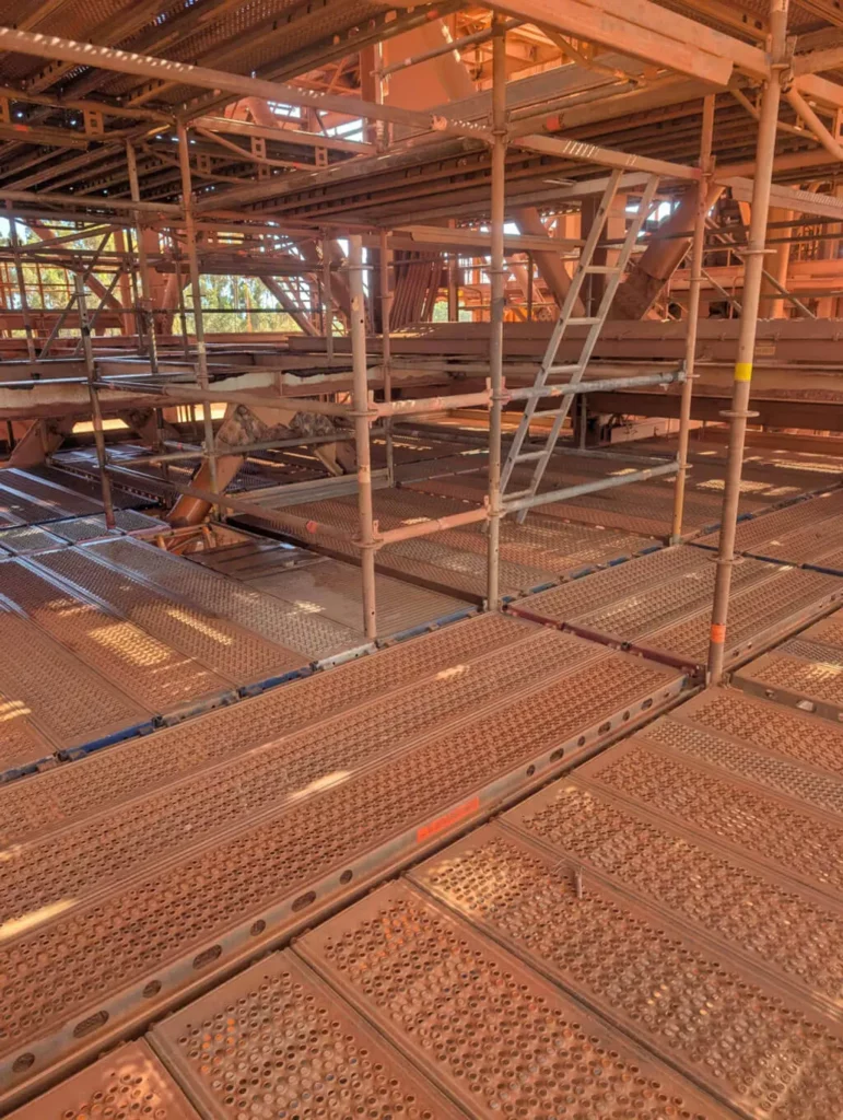 A network of metal scaffolding and grated platforms with a ladder in the center, all covered in reddish dust, possibly from mining or construction work, under bright sunlight.