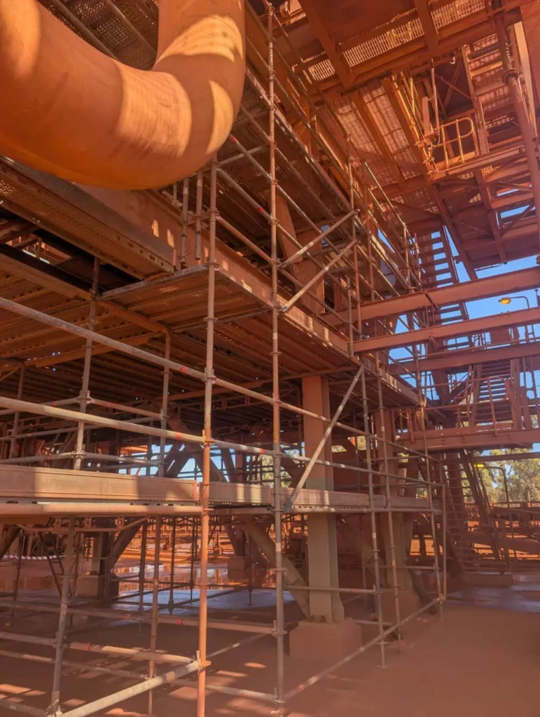 Scaffolding is set up inside an industrial facility with metal beams, stairs, and platforms. Sunlight shines through the structure, highlighting rust-colored surfaces and creating patterned shadows on the ground.
