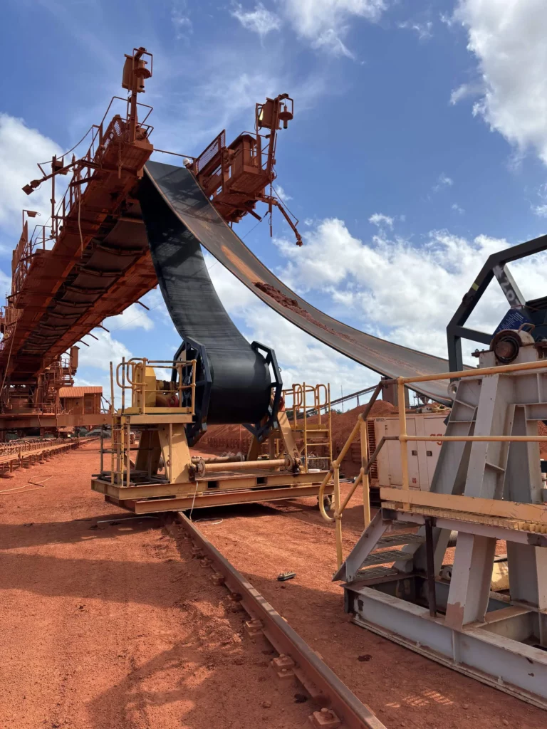A large conveyor belt installation in progress at an industrial site, with machinery and metal structures on red dirt ground under a partly cloudy sky. The conveyor belt is partially unrolled and elevated.