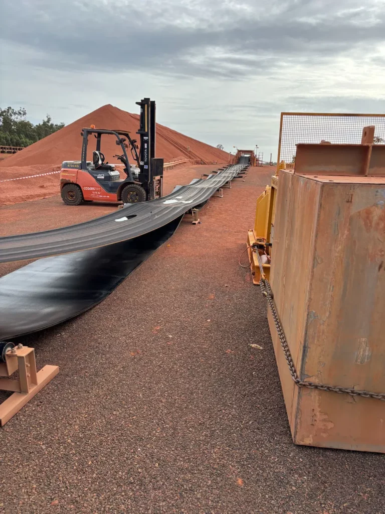 A forklift stands near a long, black conveyor belt laid out on reddish gravel, with equipment and stacked earth in the background under a cloudy sky.