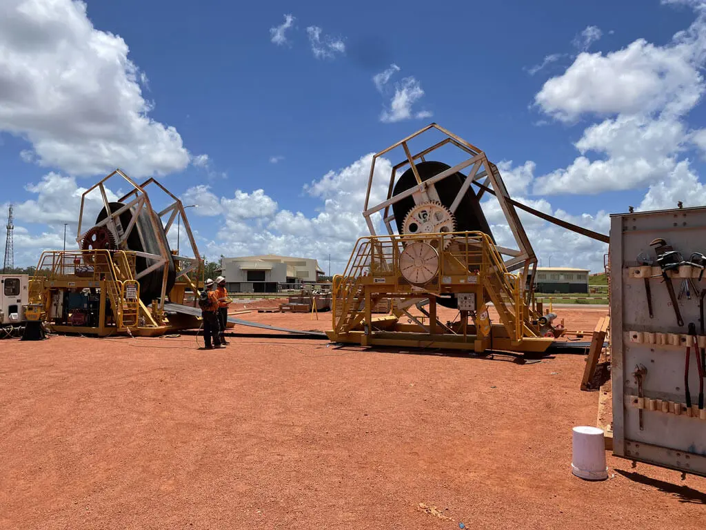 Two large industrial cable reels on yellow metal frames stand on a red dirt site under a blue sky with scattered clouds. Two workers in safety gear are nearby, with buildings and equipment in the background.