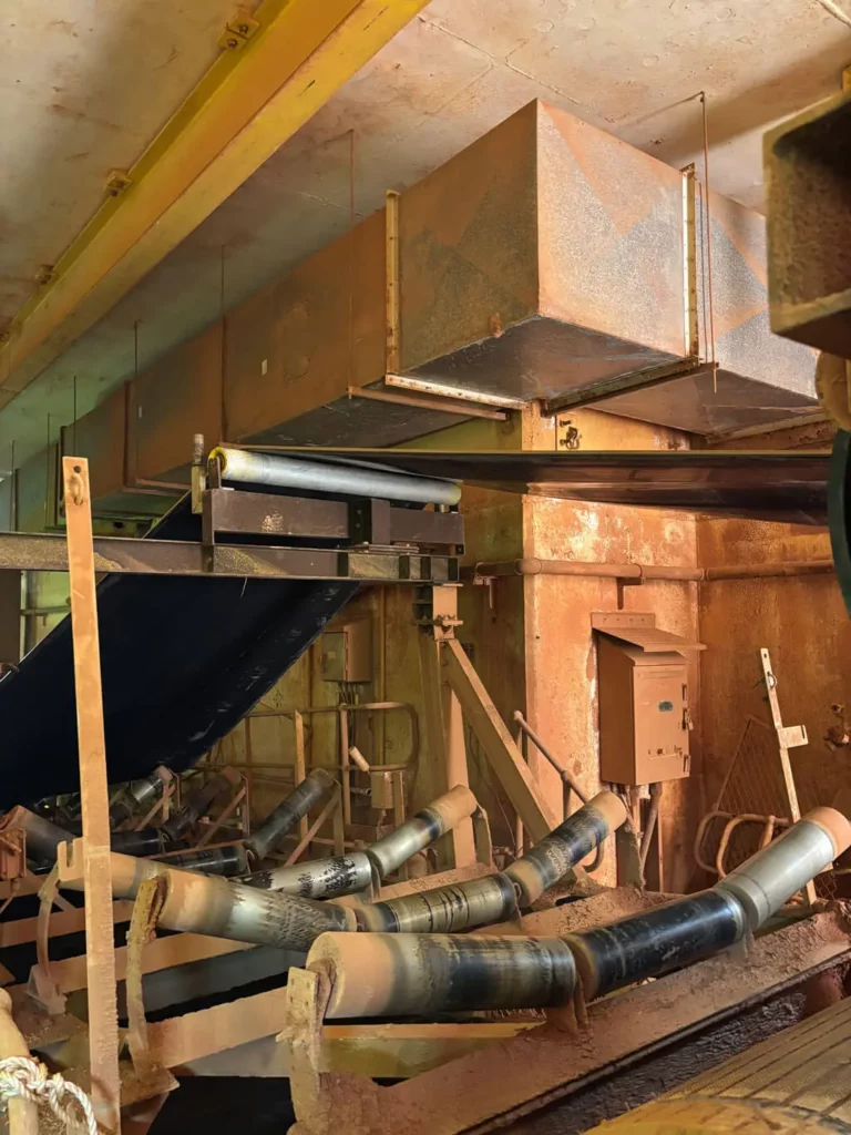 A dusty industrial conveyor system with several black rollers transports materials under a large rusty metal ventilation duct inside a factory or warehouse setting.