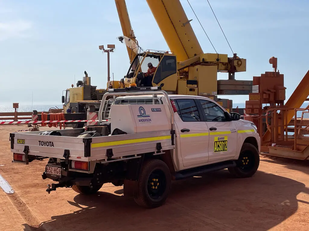A white Toyota utility truck with yellow license plates is parked on a dusty worksite near a large yellow crane. A worker in a helmet operates the crane under a clear blue sky.