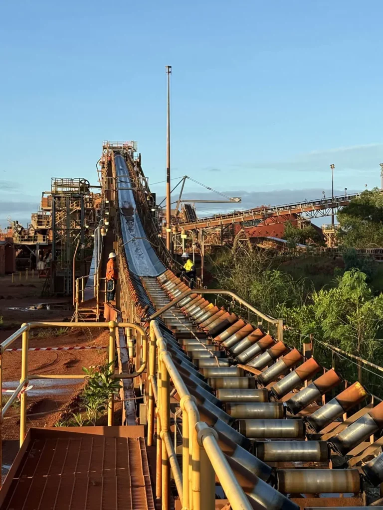 A long industrial conveyor belt transports materials at a mining site, with workers in safety gear standing nearby. The scene is surrounded by metal structures and green vegetation under a clear blue sky.