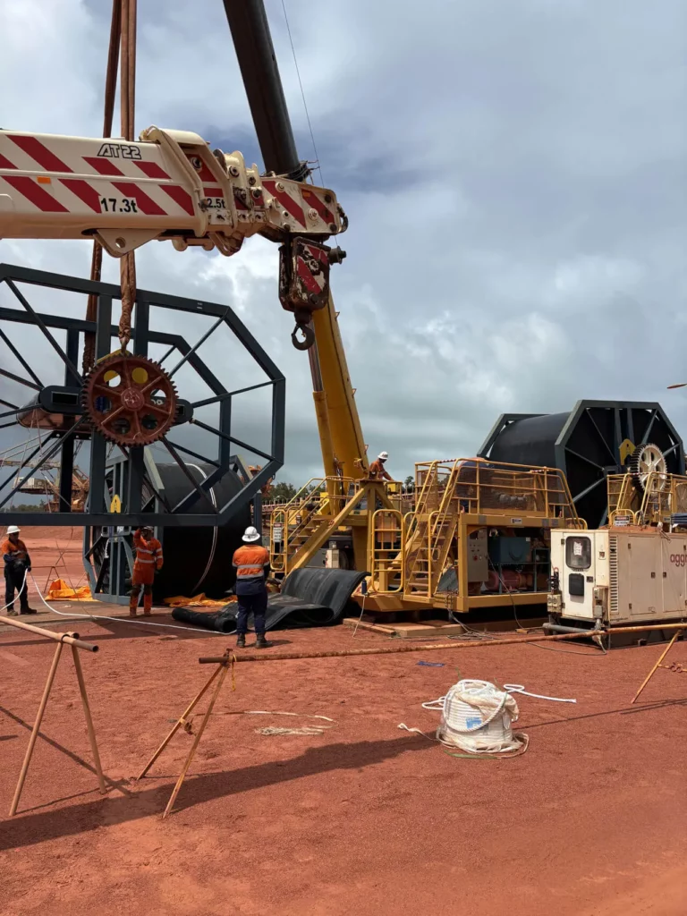 Workers in safety gear operate a large crane and heavy equipment on a red dirt construction site, moving or installing large industrial reels and machinery under a cloudy sky.