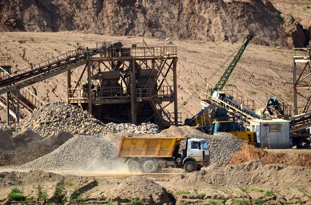 A yellow dump truck drives past piles of gravel and rocks at an open-pit mining site, with industrial equipment, conveyor belts, and cranes in the background against a rocky landscape.