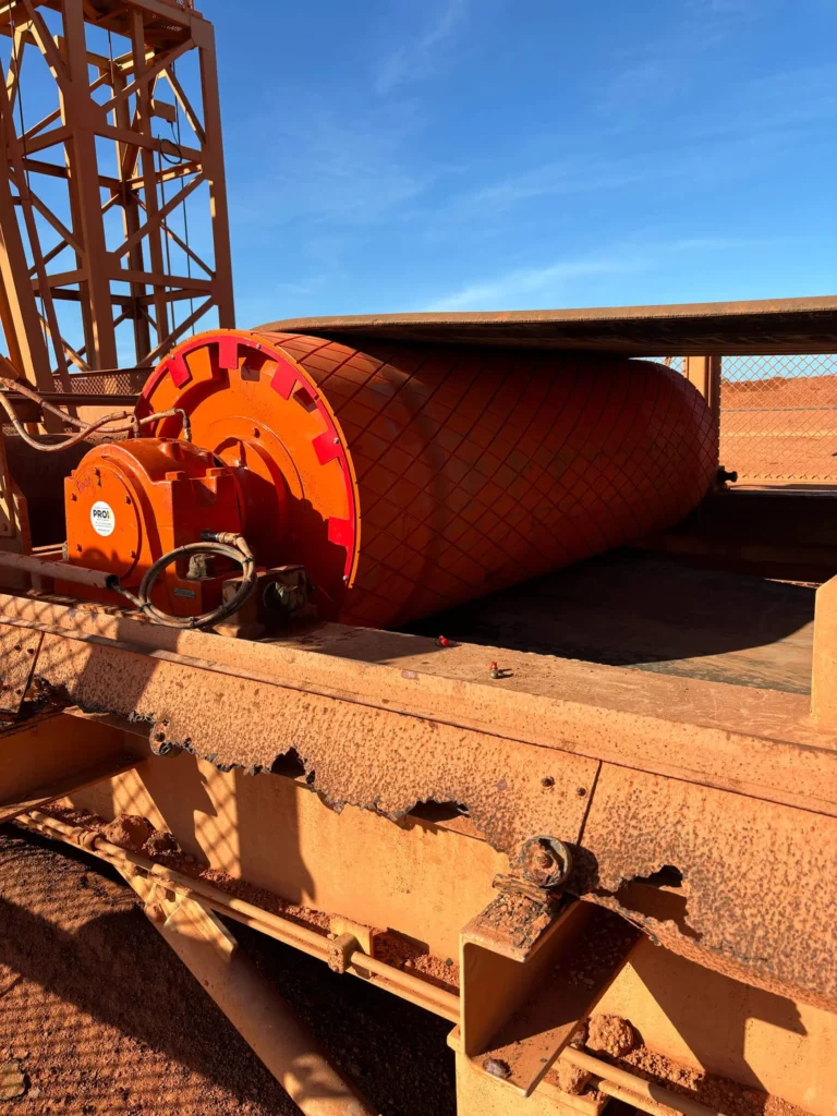 A large orange conveyor belt drum with a diamond-patterned surface sits on a rusty metal frame, under a clear blue sky in a mining or industrial setting.