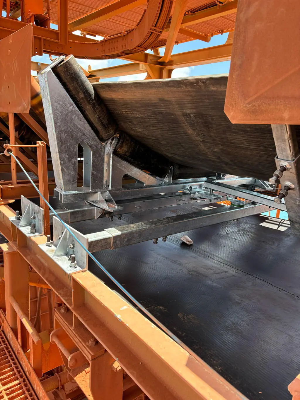 A large industrial conveyor system with metal framework and black rubber belts, used for transporting materials in a mining or manufacturing facility. Rust-colored dust and machinery are visible in the background.