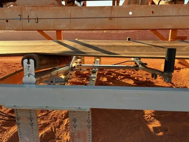 A close-up of industrial machinery, showing a conveyor belt system with metal rollers and support beams, set on red dirt ground. The machinery appears to be outdoors and is partially rusted.