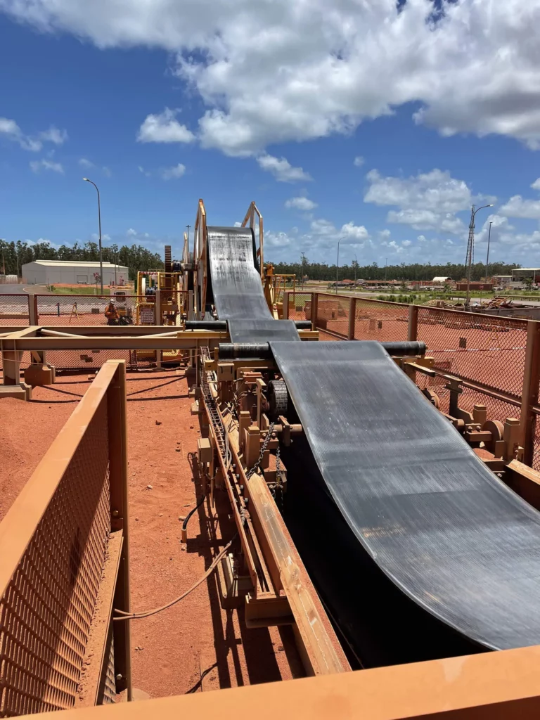 A long, black conveyor belt stretches outdoors across a reddish dirt surface, surrounded by yellow industrial frames and equipment under a partly cloudy blue sky.