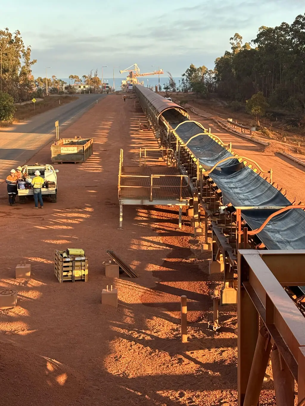 A group of workers in safety gear stands beside a pickup truck next to a long industrial conveyor belt on a dirt road, surrounded by trees and construction equipment under a partly cloudy sky.