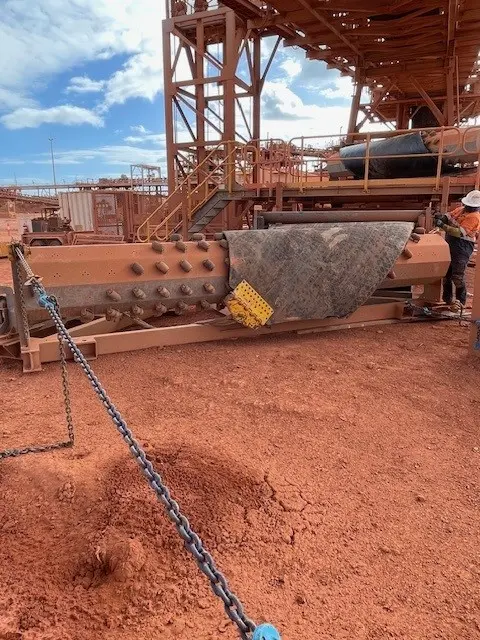 A large industrial machine sits on red dirt at a mining site, partially covered with a black mat and yellow safety guard. Metal structures, stairs, and a worker in orange safety gear are visible under a blue sky.
